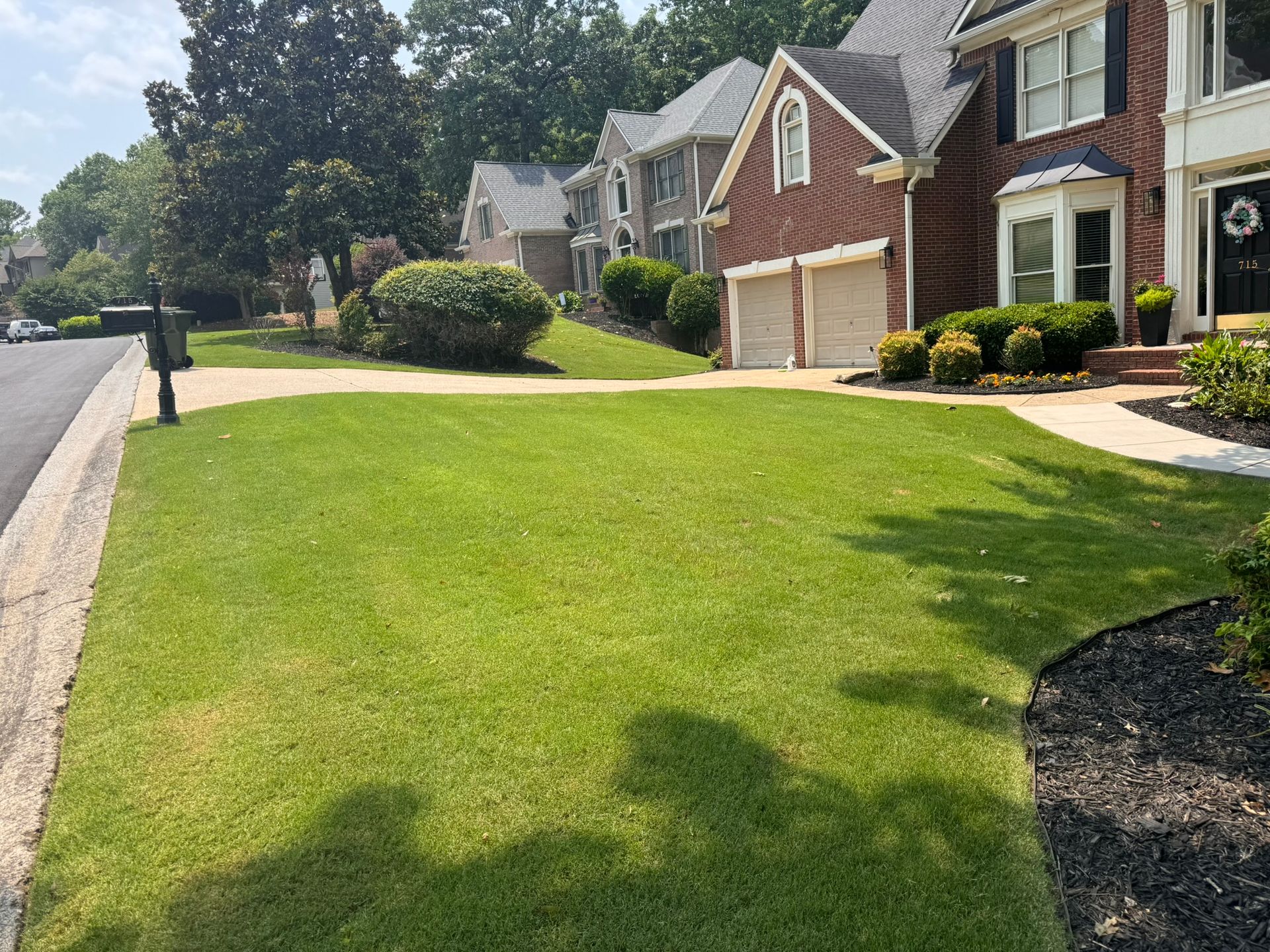 Suburban homes with manicured green lawn, driveway, and trees on a sunny street
