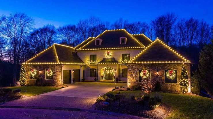 Woodstock House decorated with warm white Christmas lights at dusk, with lit shrubs and a driveway.