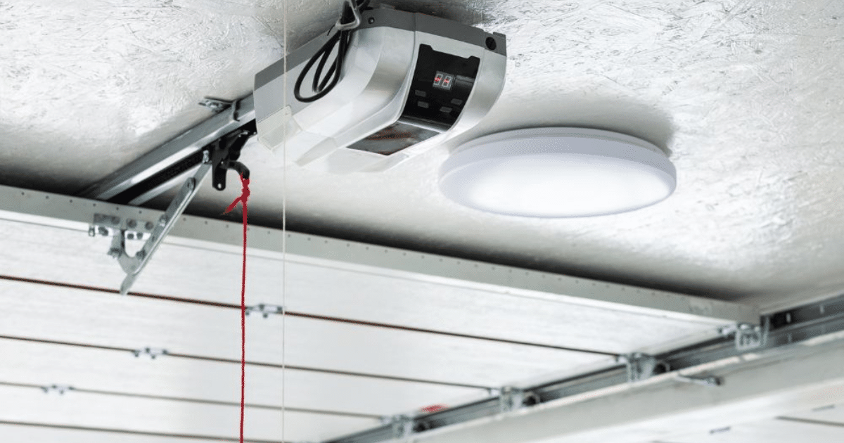 Garage door opener and attached track mounted to a white ceiling, with a white, circular light fixture nearby.