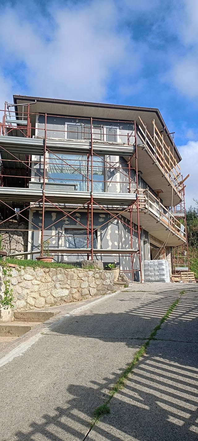 Casa in pietra con impalcature metalliche rosse e un sentiero in pendenza sotto un cielo azzurro