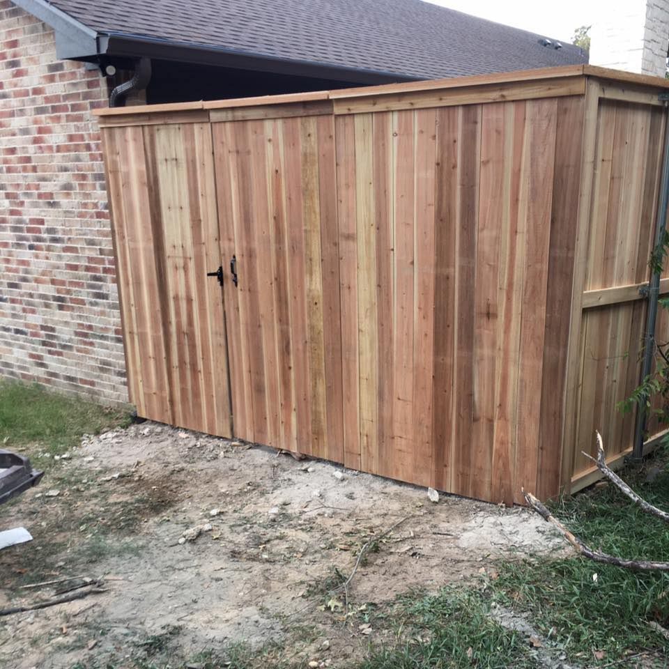 Wooden storage shed next to a brick wall and a house with a brown roof.