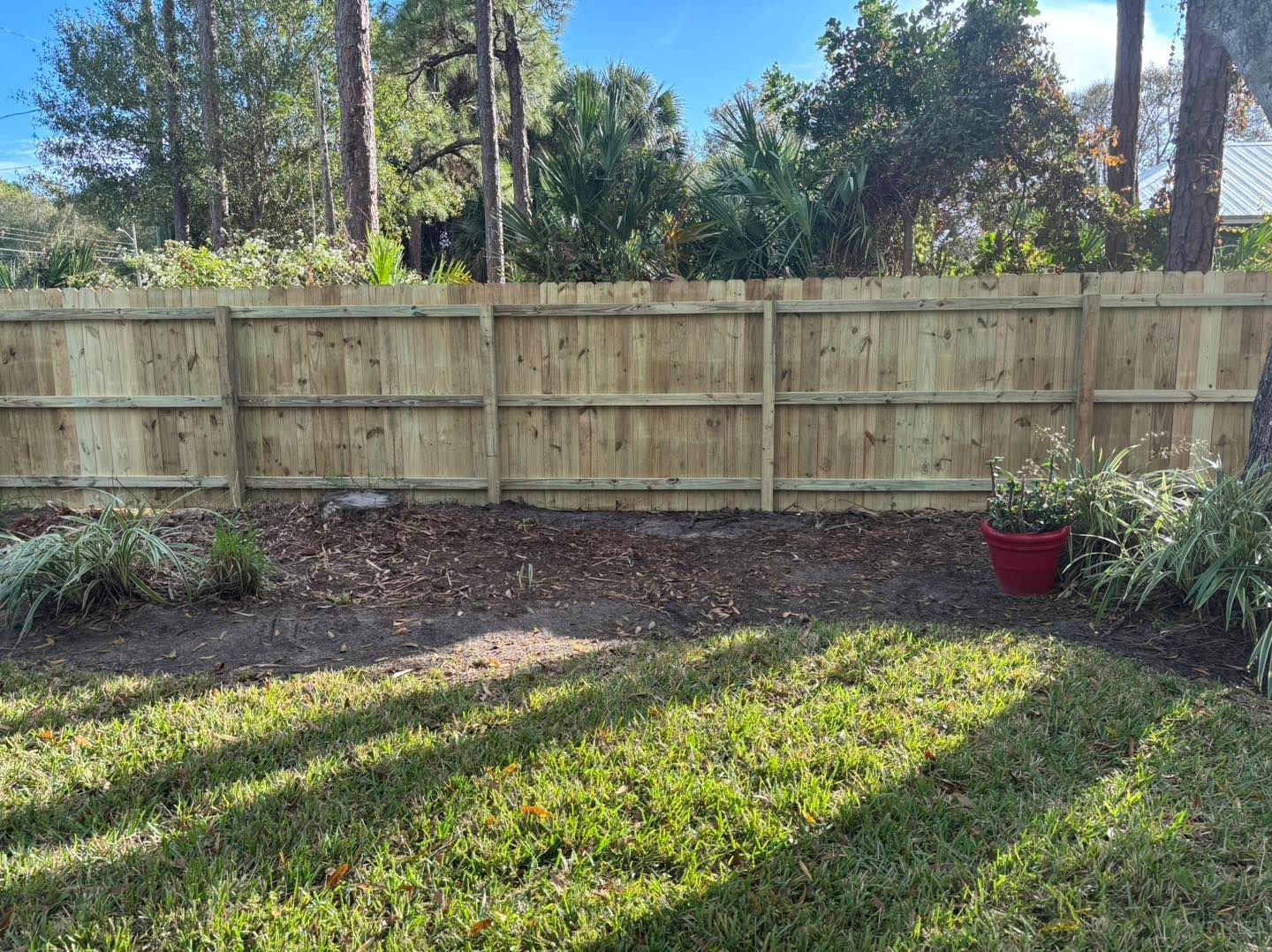 Wooden fence in backyard with plants, red pot, and green grass.