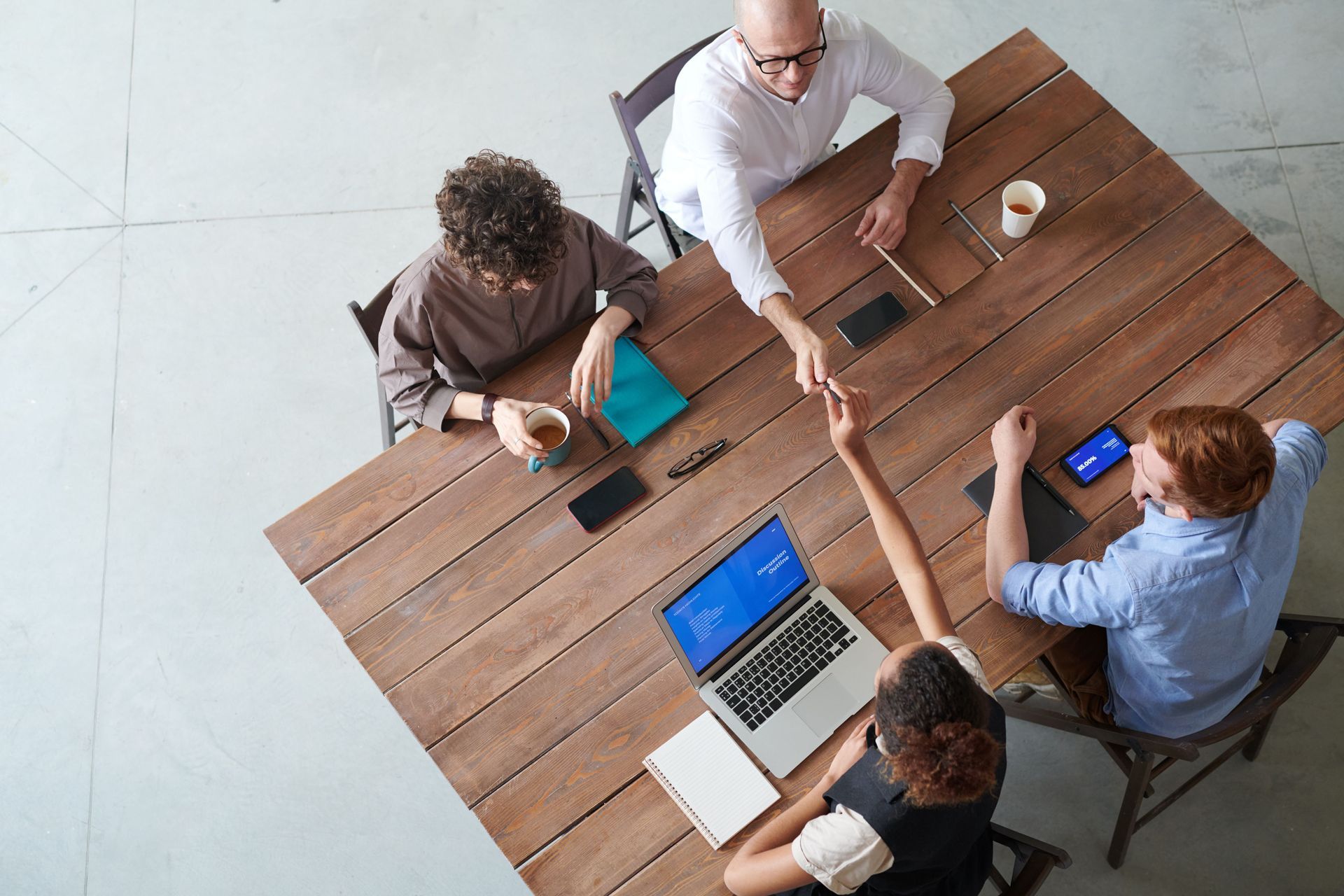 Four people collaborating around a wooden table with laptops and a smartphone in a bright office.