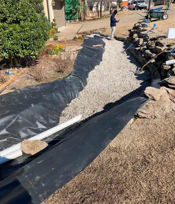 Partially installed gravel pathway lined with black tarp and rocks, with a person standing nearby