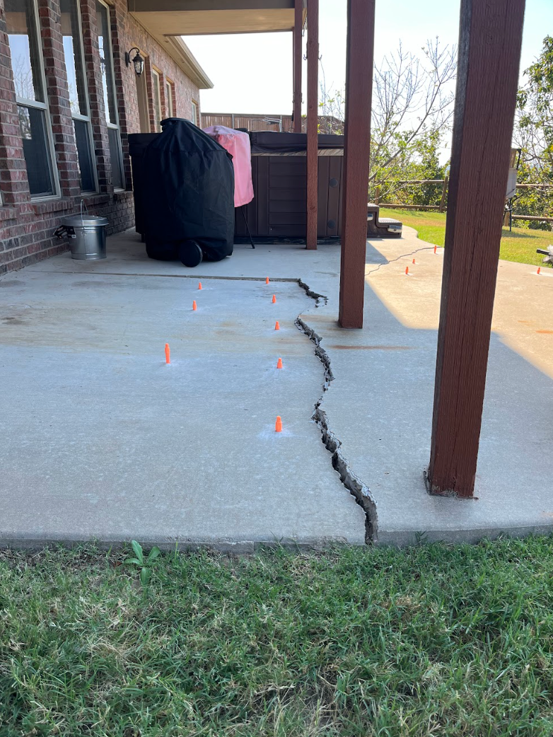 Cracked concrete porch with orange markers beside a house and wooden post, near a yard.