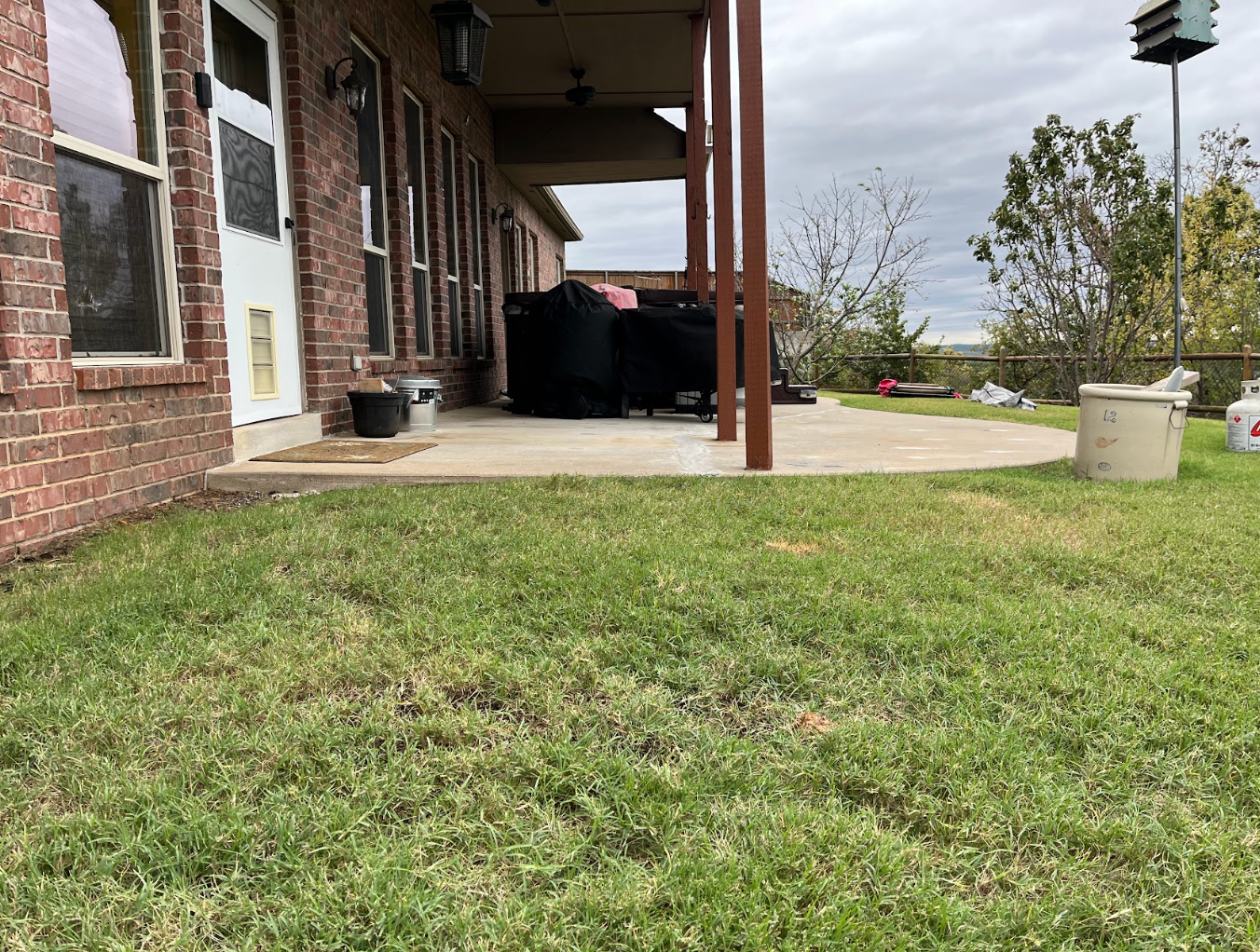 Brick porch with concrete patio, grassy yard, columns, and a bird feeder on a cloudy day