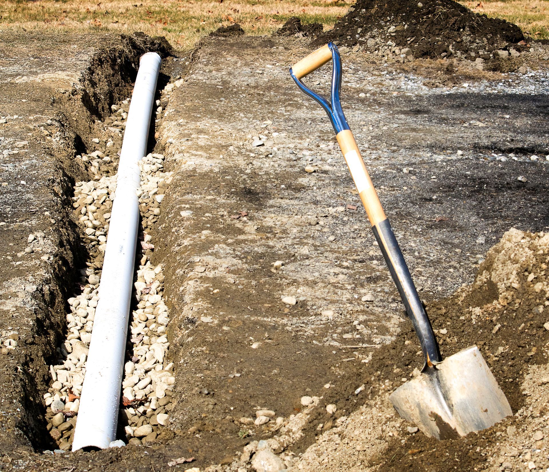 Shovel beside a freshly dug trench with a white pipe laid in gravel