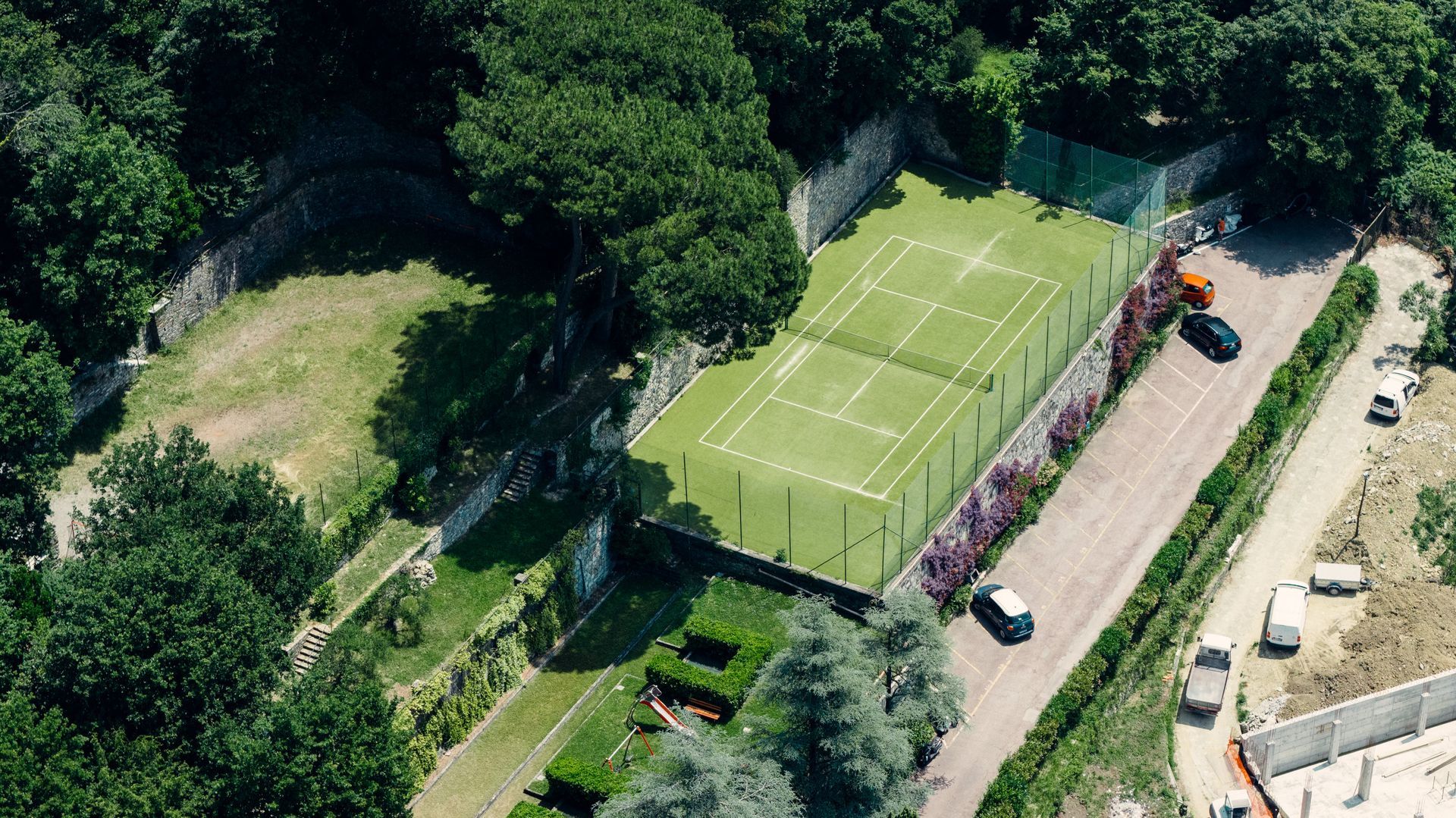 Aerial view of a green tennis court beside a dirt road, trees, and parked vehicles.
