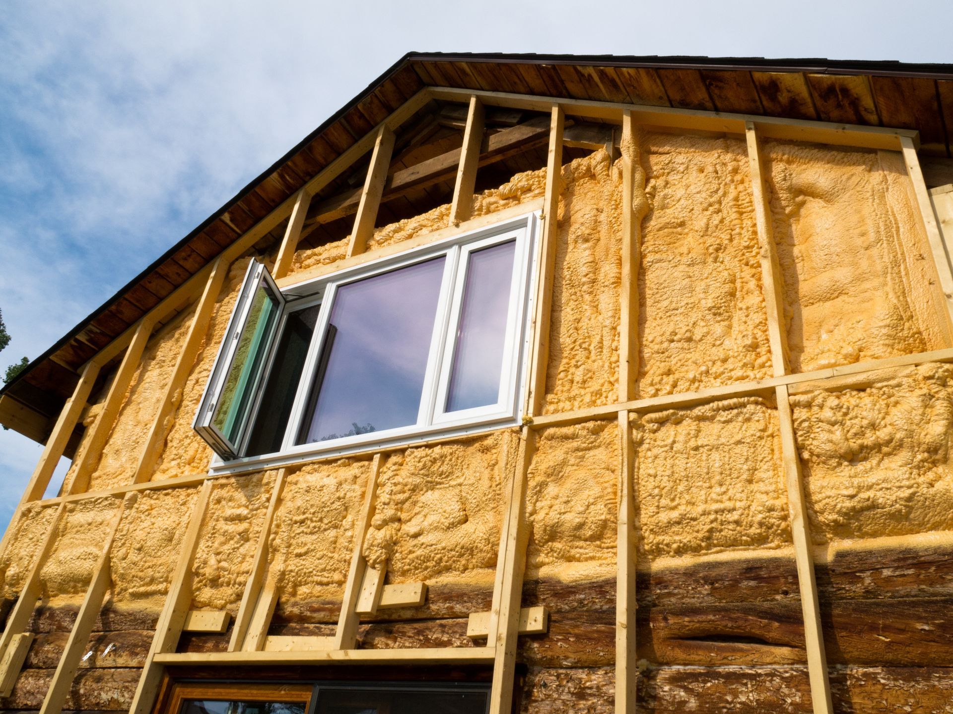 Partially built house exterior with exposed wood framing, yellow insulation, and a window under a blue sky