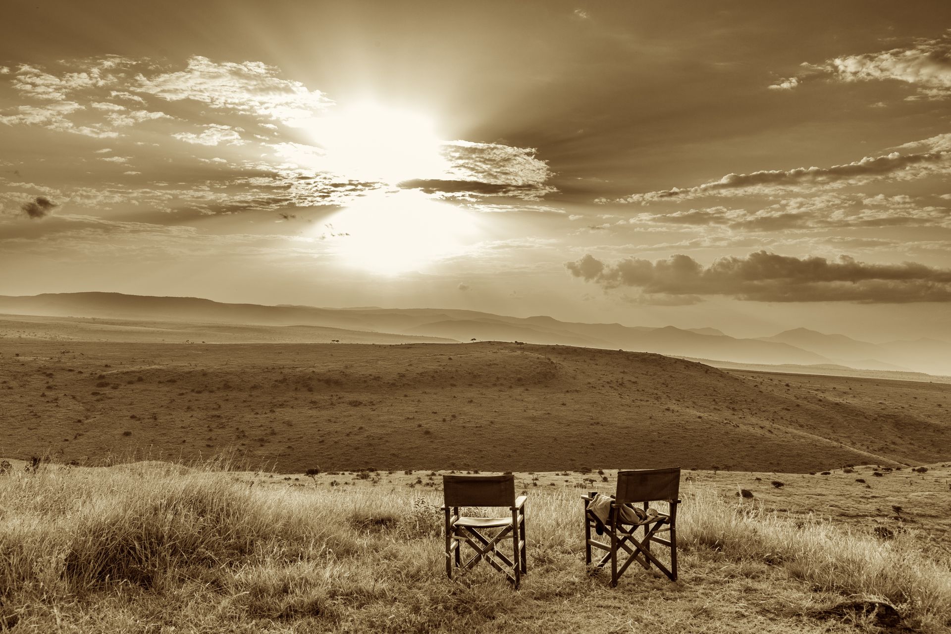 Two empty chairs facing a sepia sunset over rolling hills and a grassy field