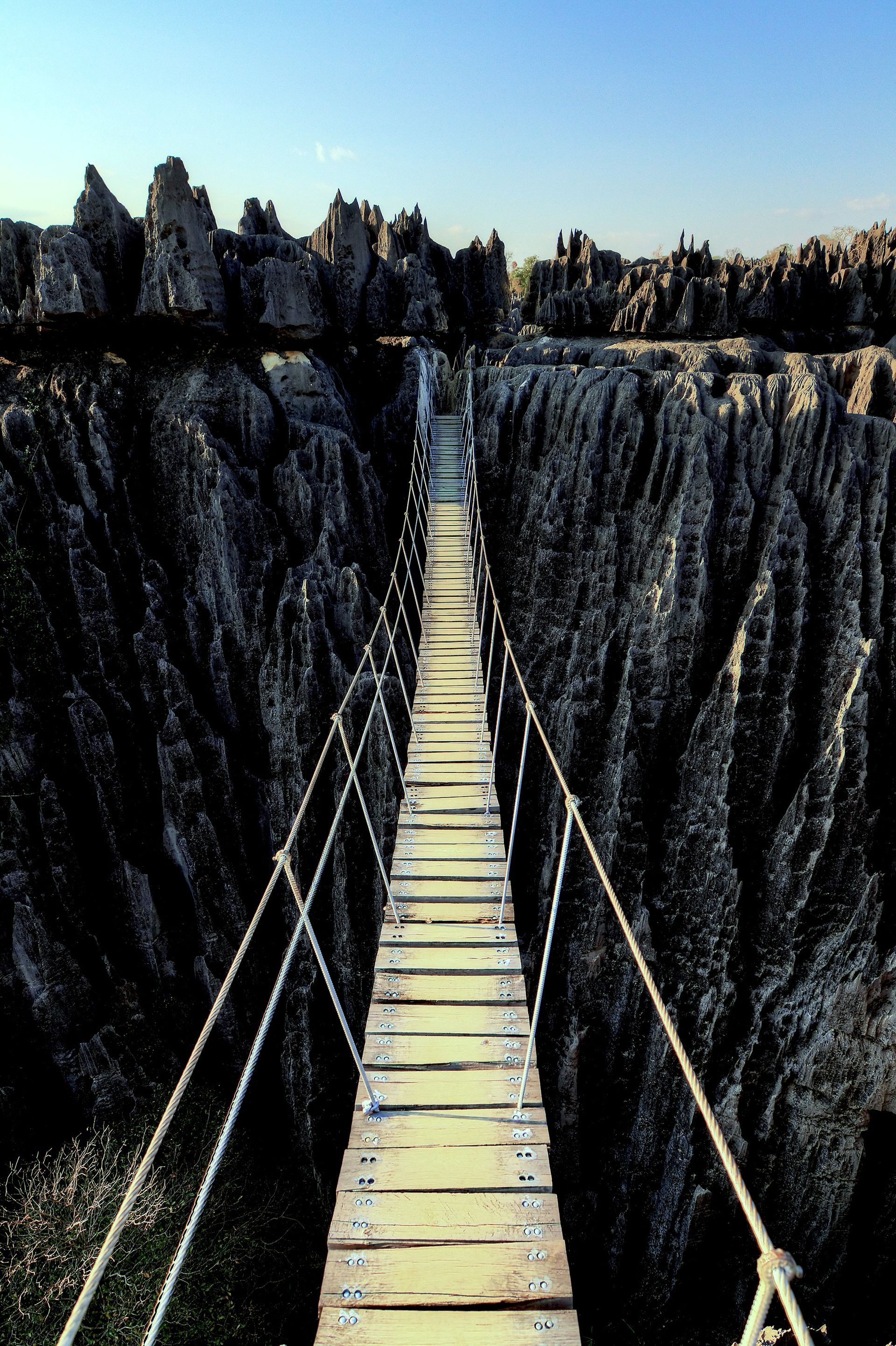 Narrow wooden bridge over a deep rocky chasm under a clear blue sky