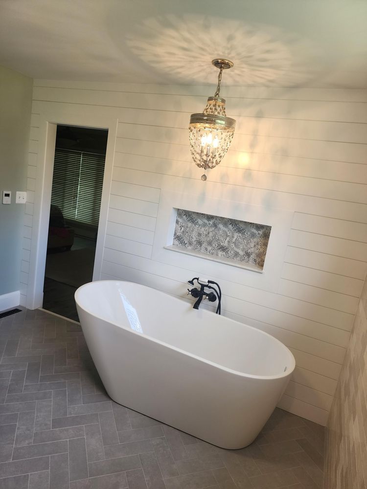 Bathroom with white tub, shiplap wall, stone niche, and ornate chandelier. Herringbone floor.