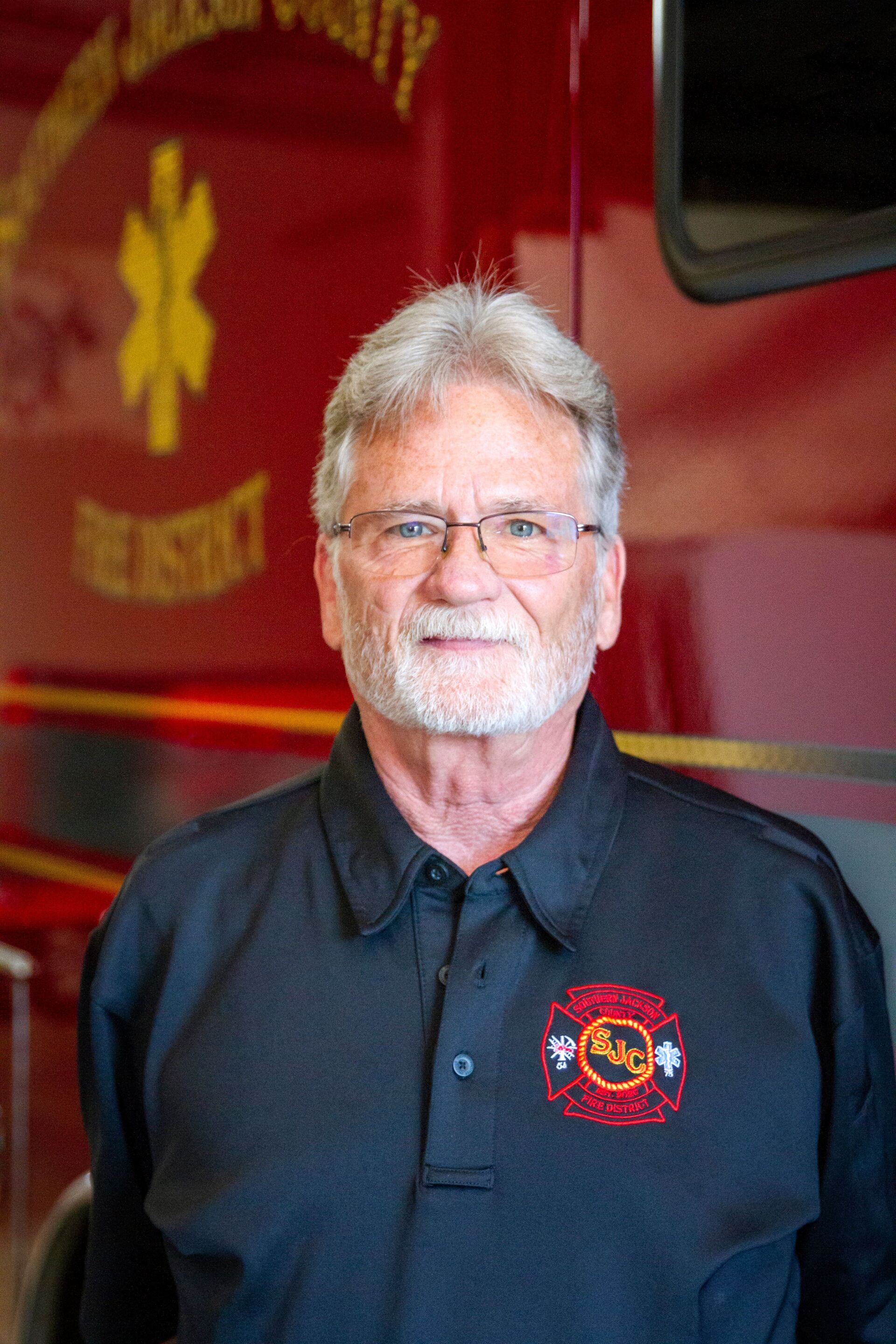 Smiling man in navy polo with embroidered logo, standing in front of a red fire truck.