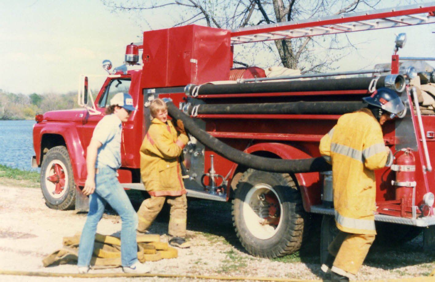 Workers in yellow rain gear load a red fire truck with black hoses outdoors