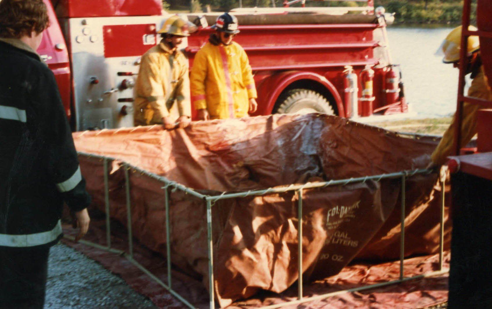 Firefighters beside a red fire truck inspect a large brown tarp-covered container at a roadside scene
