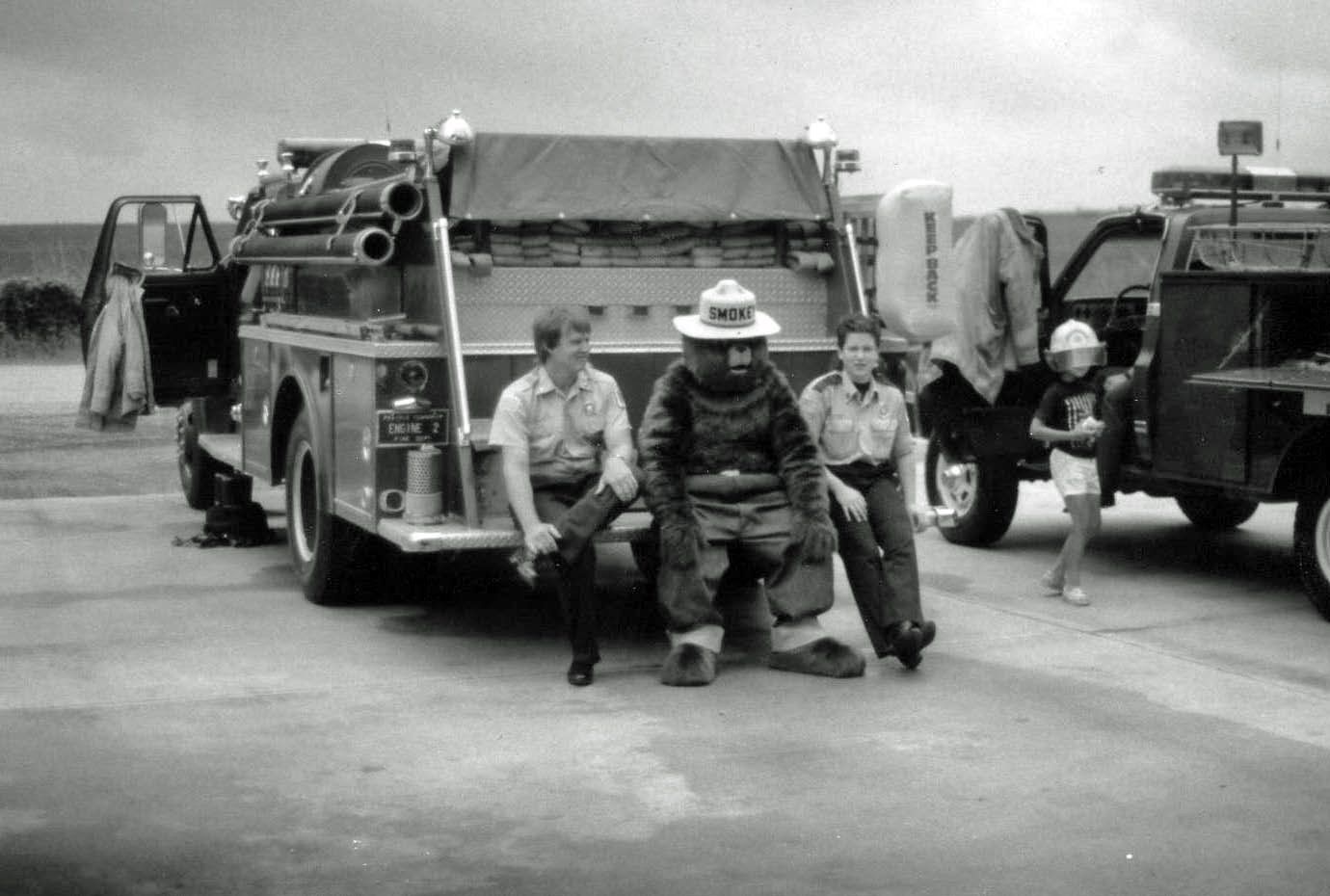 Firefighters posing in front of a fire truck on a road, with equipment and vehicles behind them