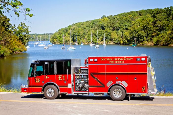 Red fire truck parked by a lakeside road with sailboats and green hills in the background