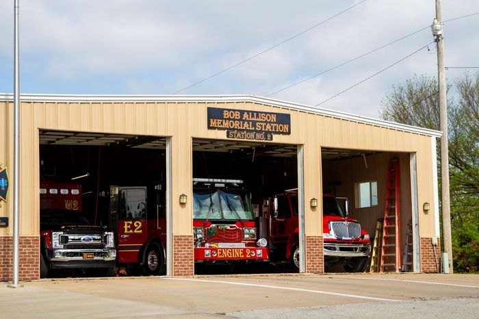 Fire station with three open bays and fire trucks parked inside, under a beige roof.