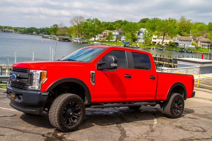 Red lifted pickup truck parked by a waterfront with houses and trees in the background