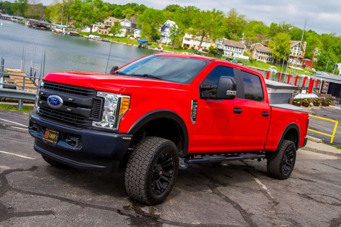 Red lifted Ford pickup truck parked by a waterfront road, side view.