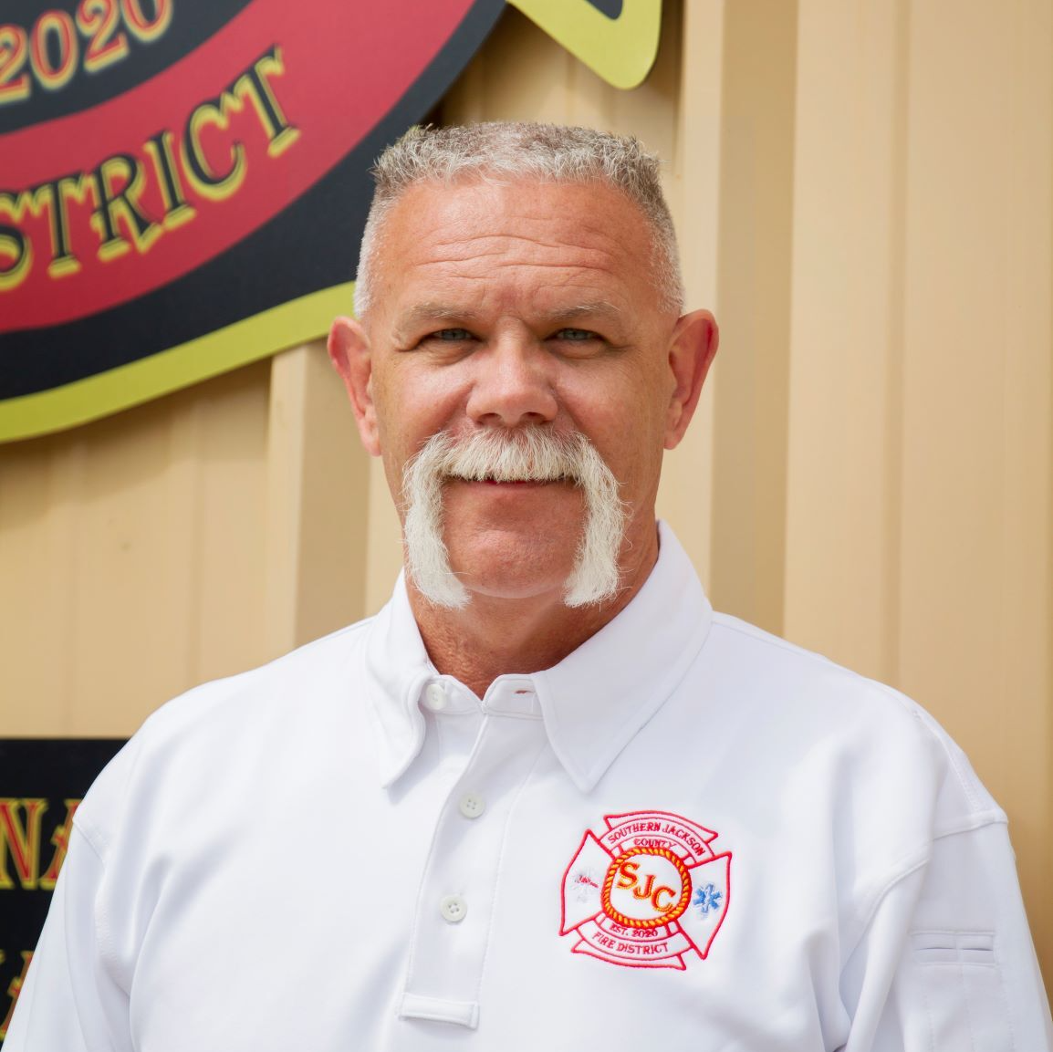 Man in white polo standing before a beige wall with a colorful district emblem