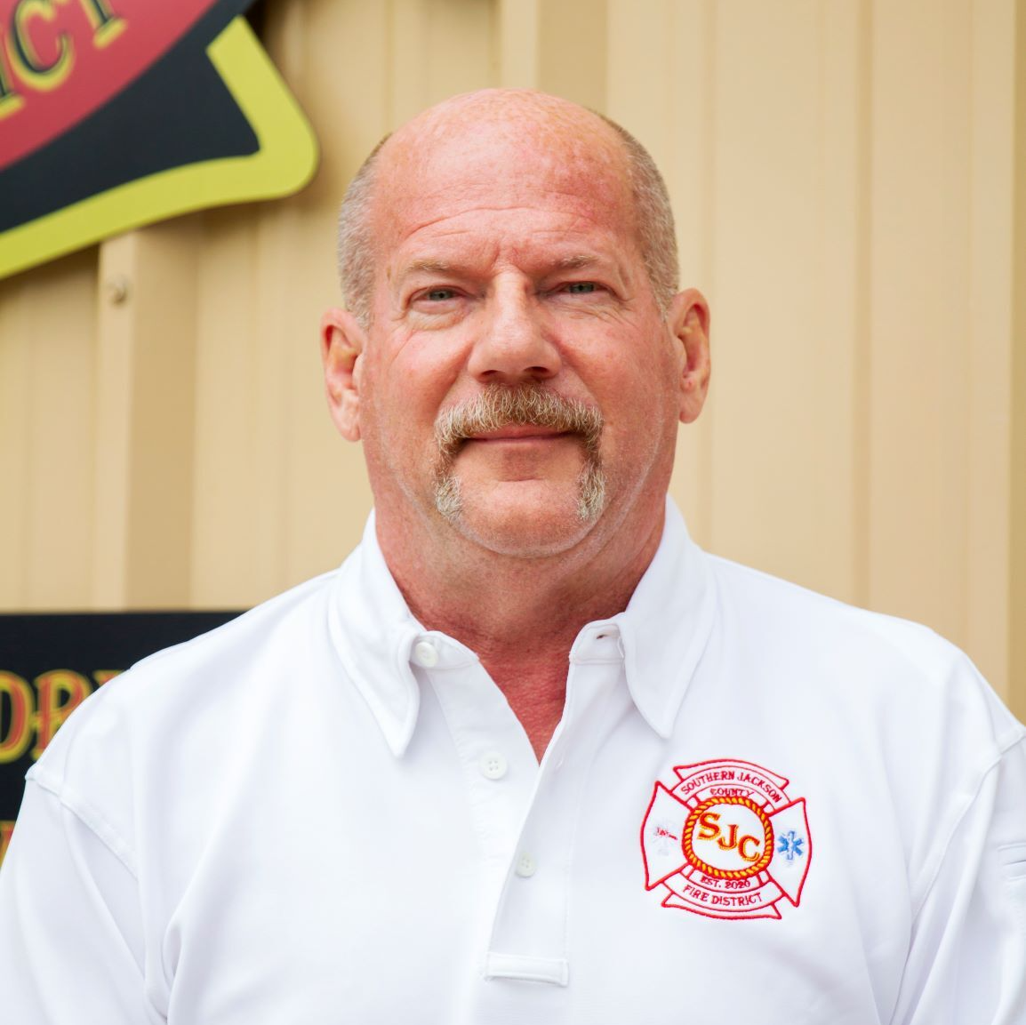 Bald man in a white polo shirt standing indoors against a beige wall.