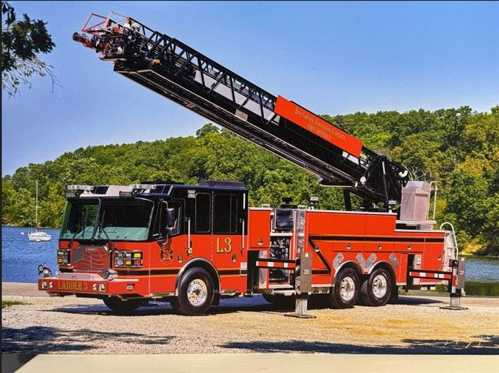 Red fire truck with extended ladder parked on a sandy shore near water and trees
