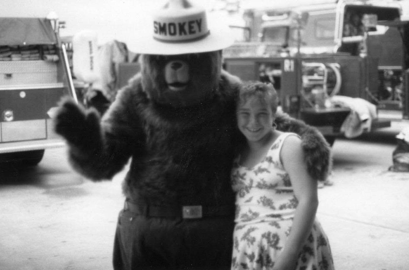 Person in a Smokey Bear costume posing with a smiling girl in front of trucks