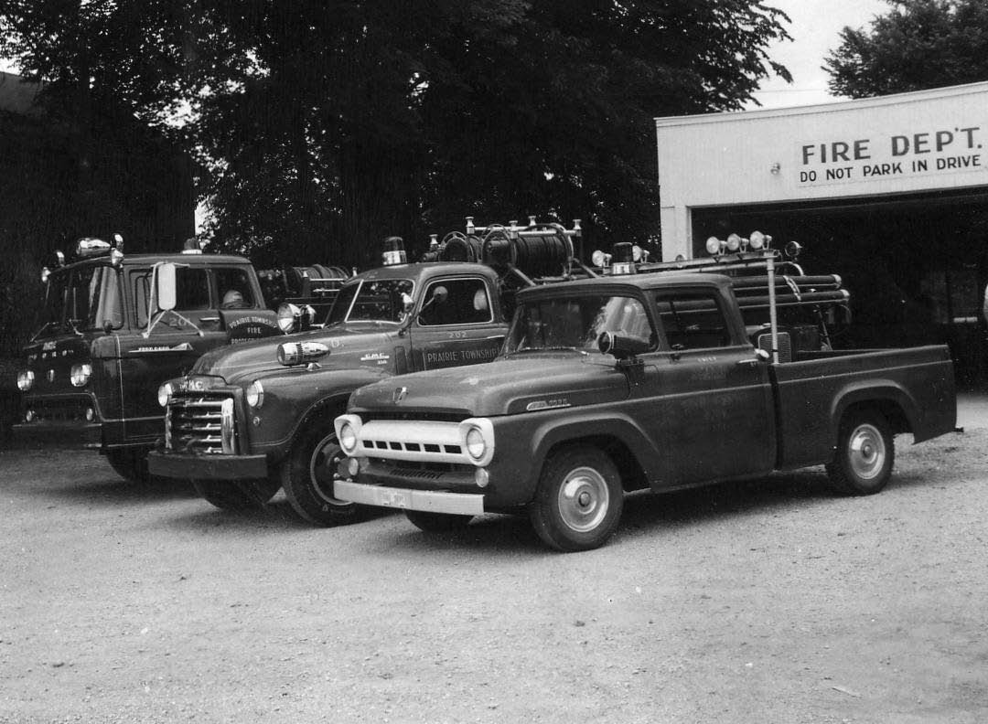 Vintage fire department trucks parked in front of a fire station building.