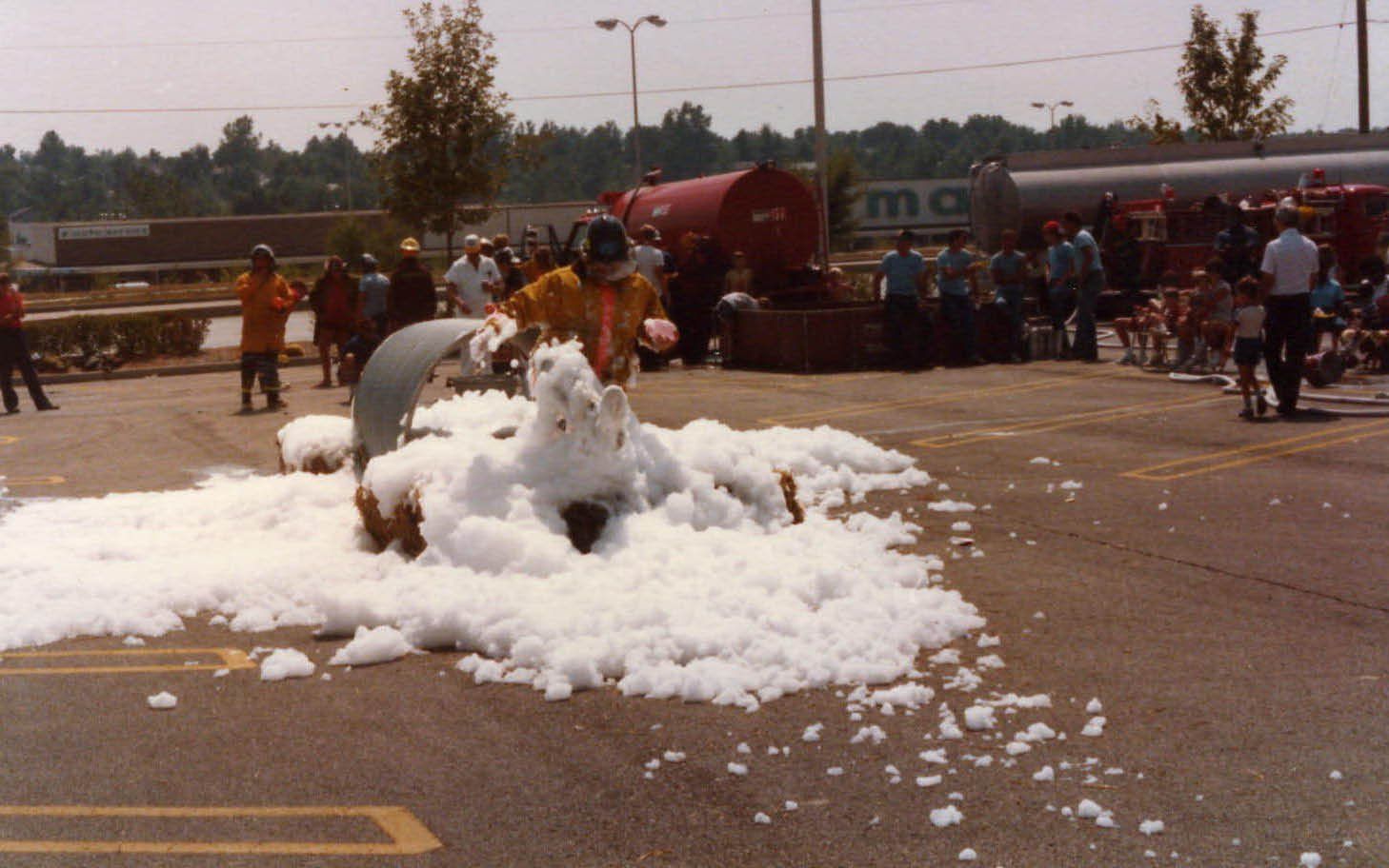 A white foam pile on pavement at an outdoor event, with people and a fire truck in the background.