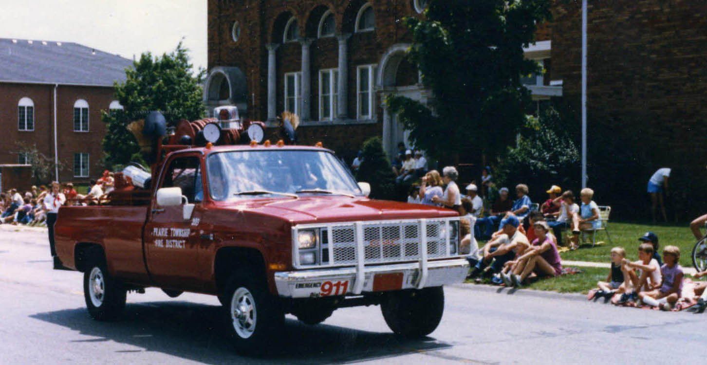 Red emergency pickup truck driving in a parade with passengers and spectators watching