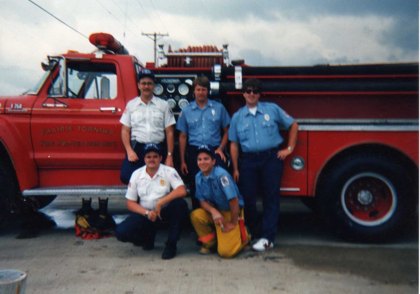 Five firefighters posing beside a red fire truck in front of a cloudy sky.