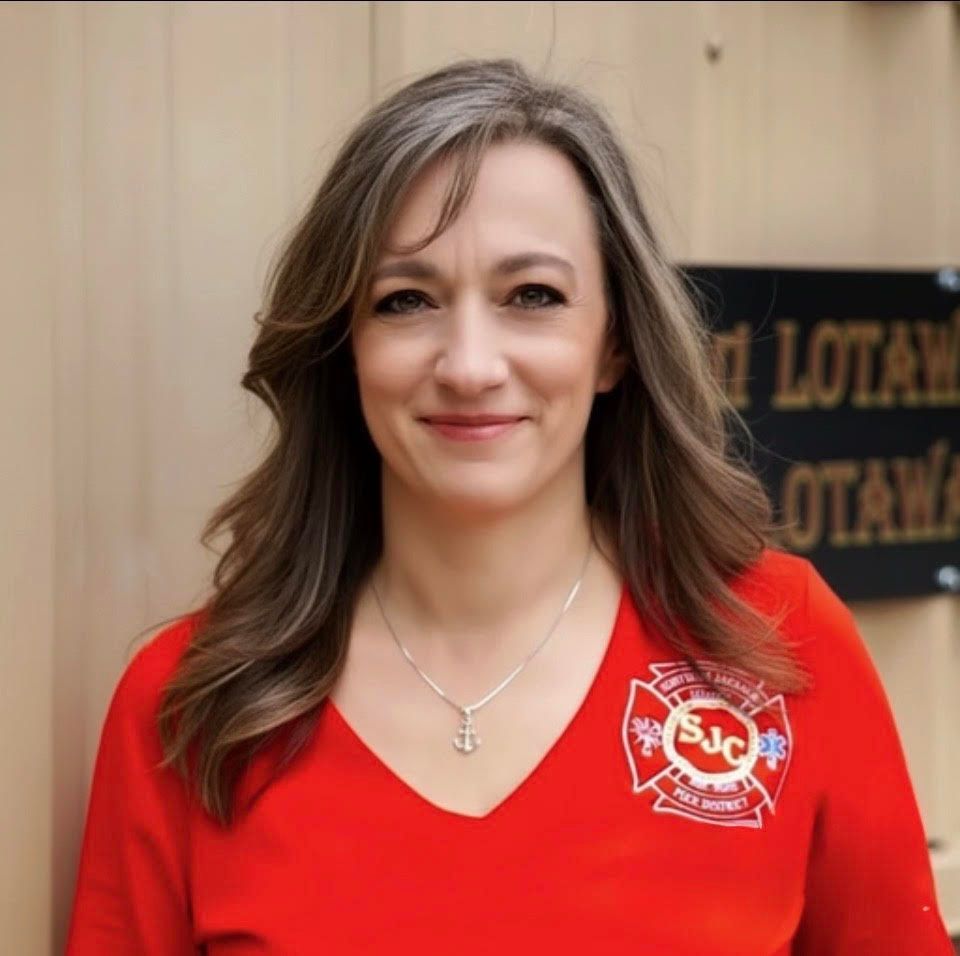 Woman in red shirt with a white emblem, smiling against a wooden wall and dark sign.