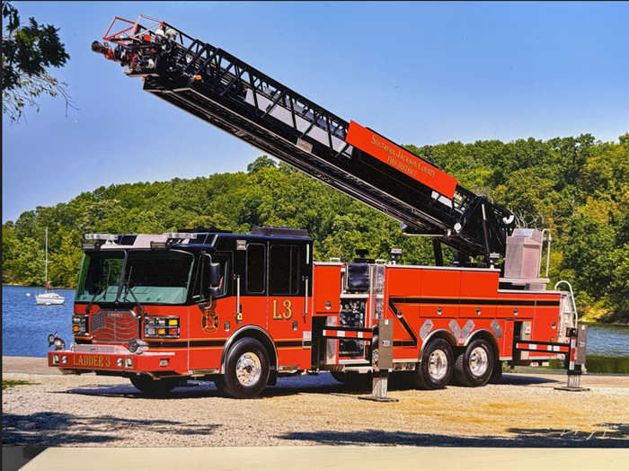 Red fire truck with raised ladder parked by a lake and trees on a sunny day