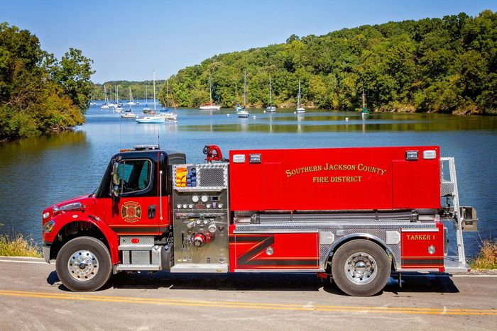 Red fire truck parked by a river with sailboats and trees in the background