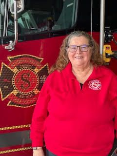 Woman in a red shirt standing beside a red fire truck with a fire department emblem.