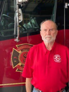 Man in a red polo shirt standing in front of a fire truck door with a fire department emblem.
