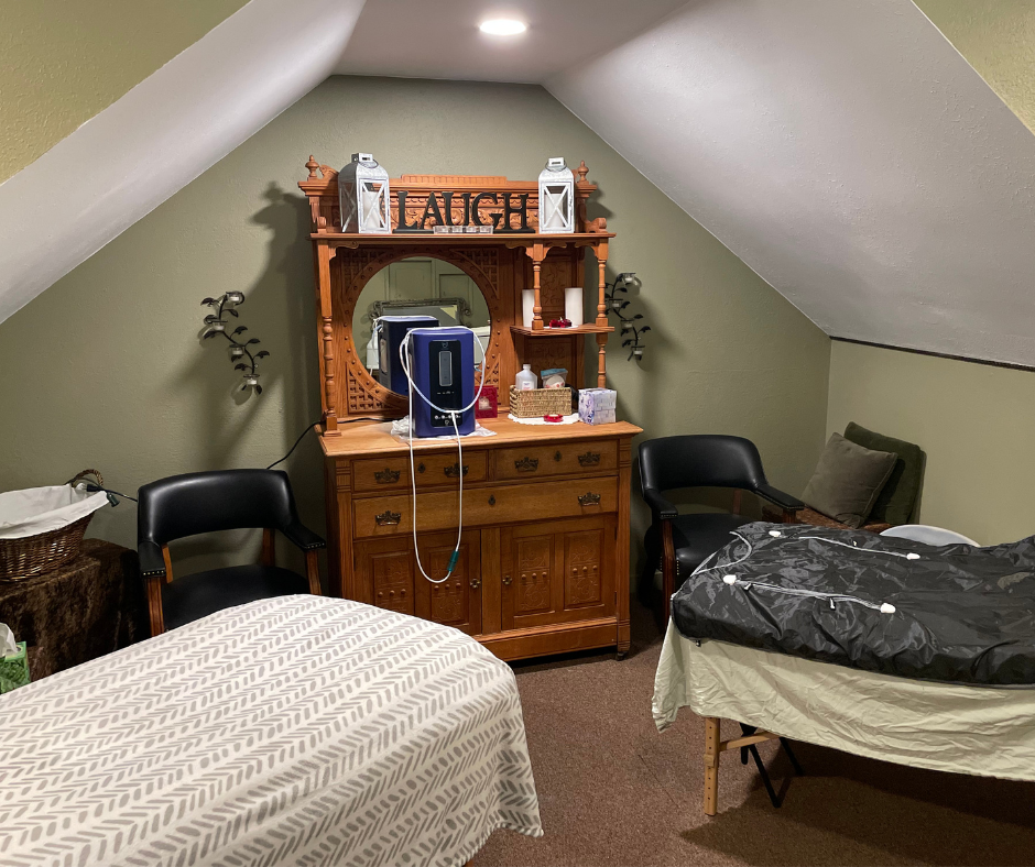 A wellness room with two massage tables, two chairs, and a wooden vanity under a sloped ceiling painted olive green.