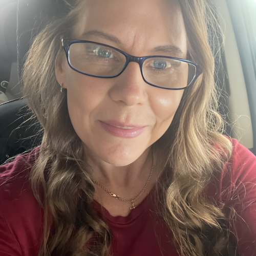 A person with wavy blonde hair wearing glasses and a maroon top, posing for a selfie inside a vehicle.