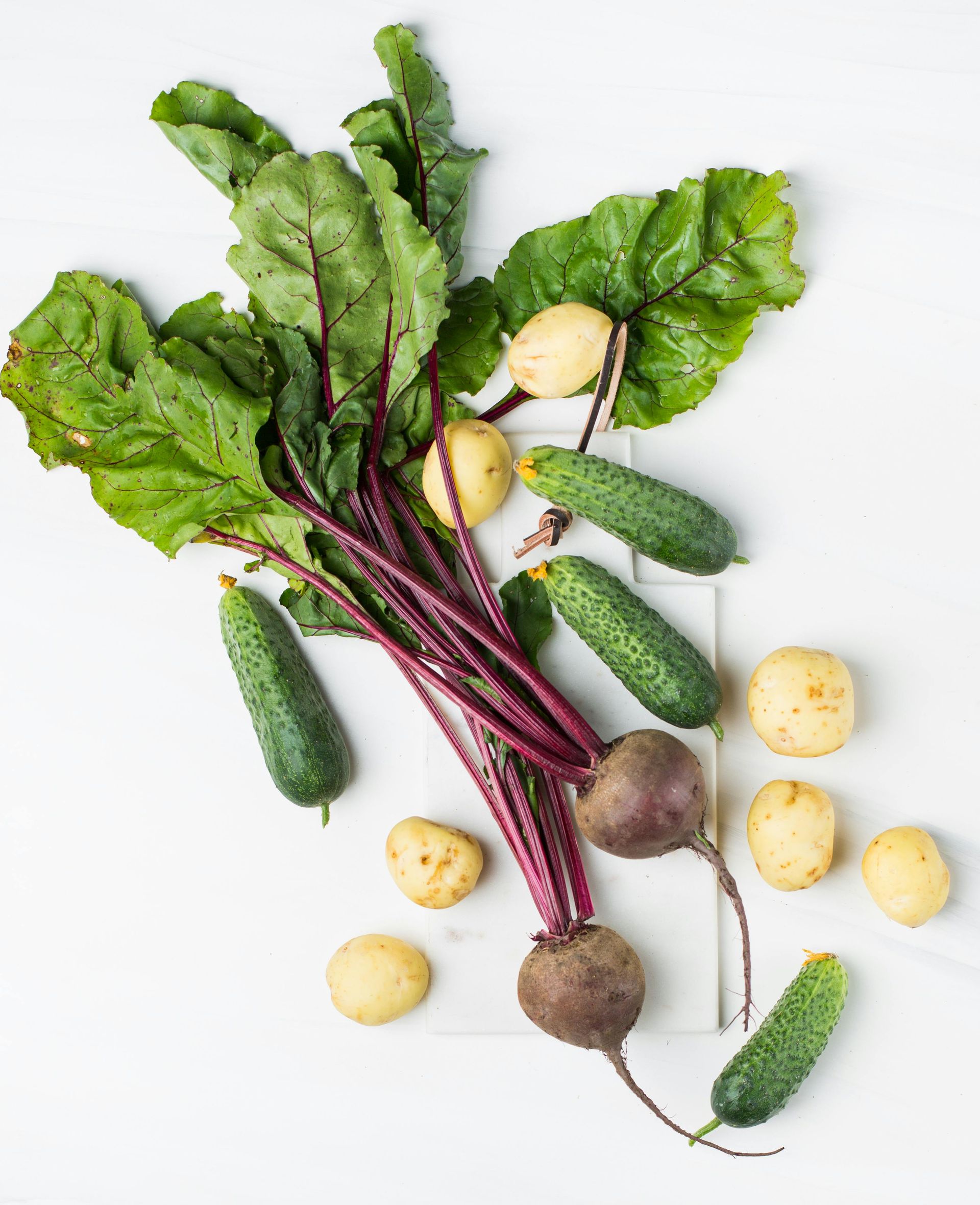 Fresh beets with leafy greens, small cucumbers, and yellow potatoes arranged on a plain white surface.