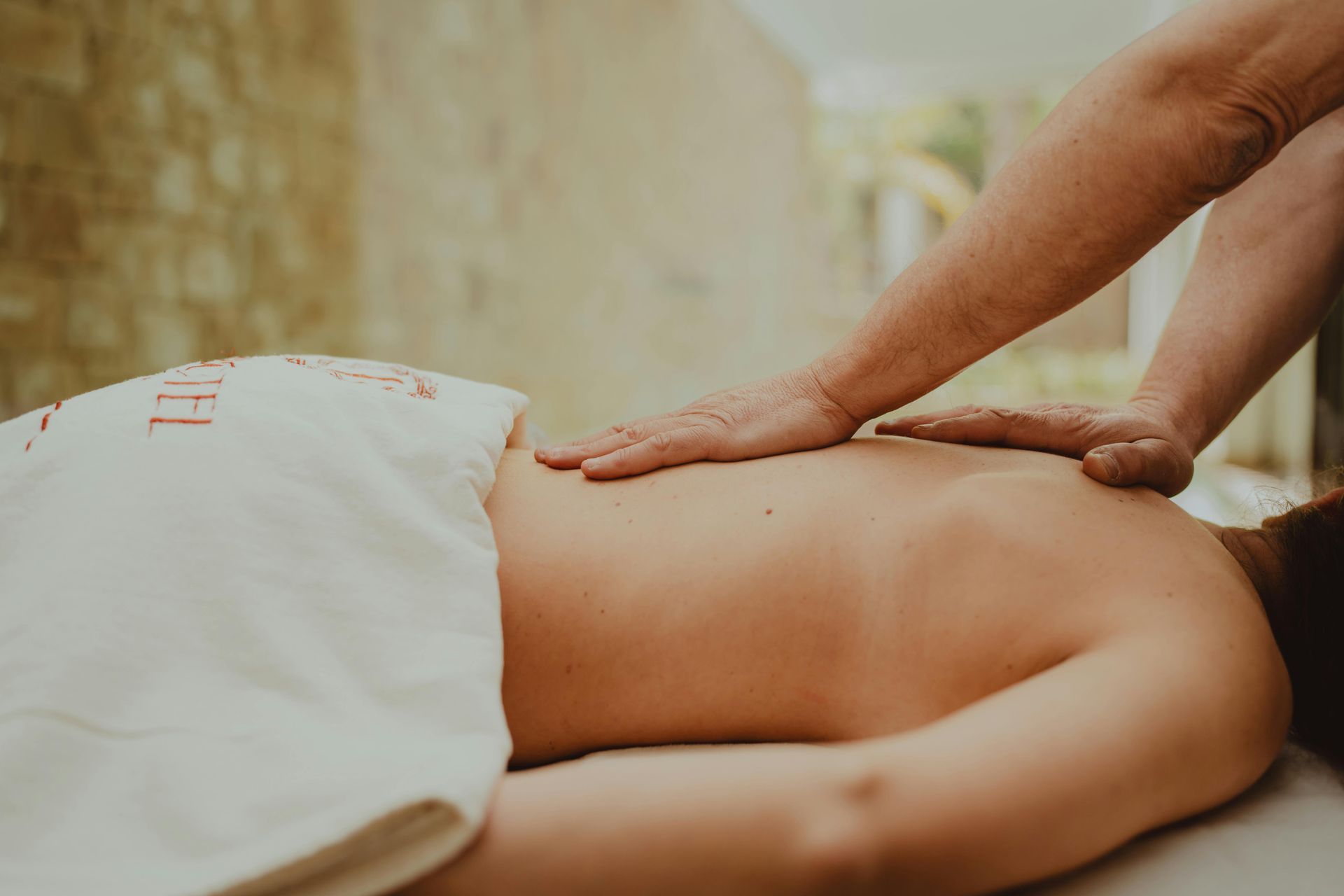 A person receives a back massage in a spa setting, with hands pressing firmly on their upper back.