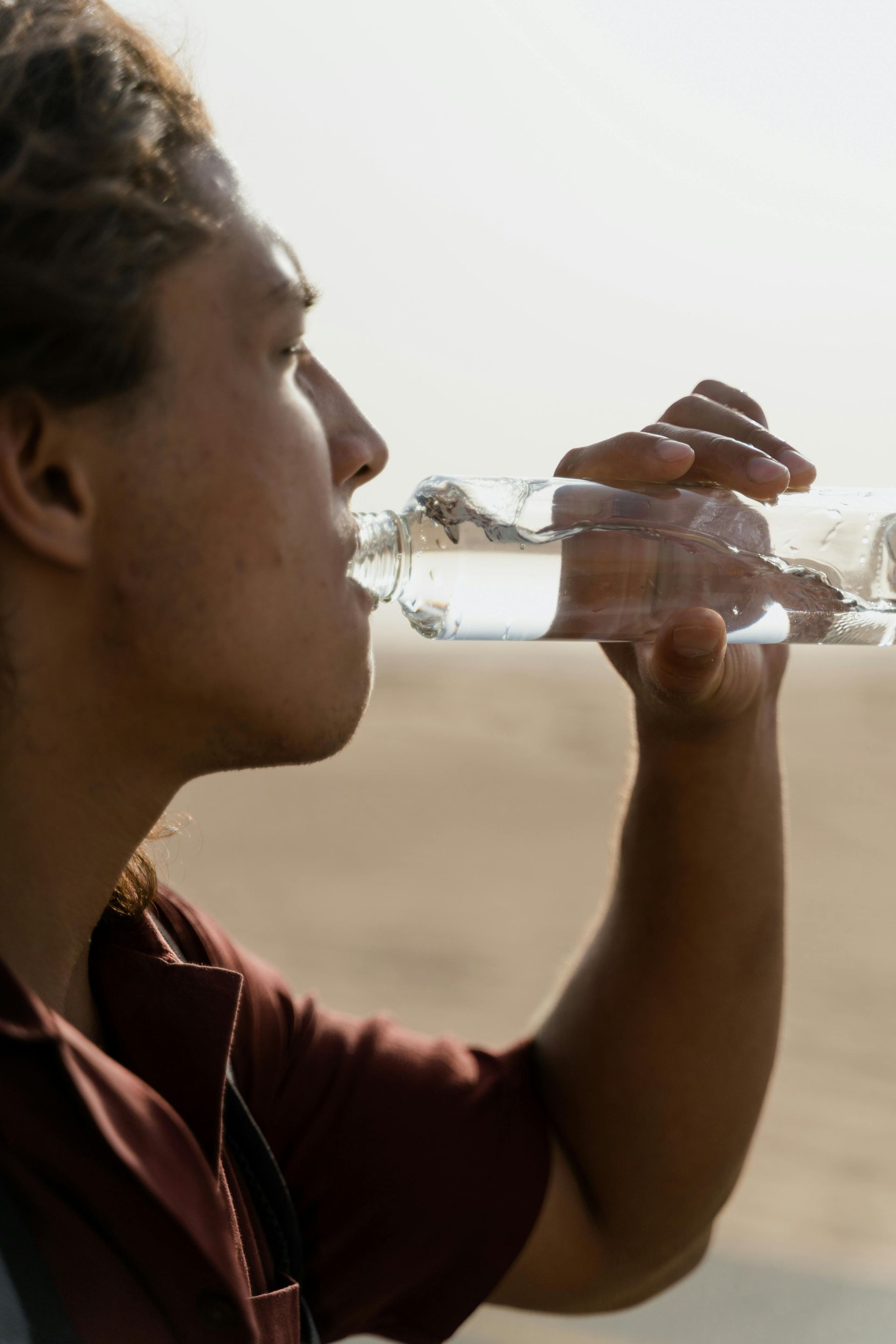 A person in a maroon shirt drinks from a clear plastic water bottle against a sandy outdoor background.