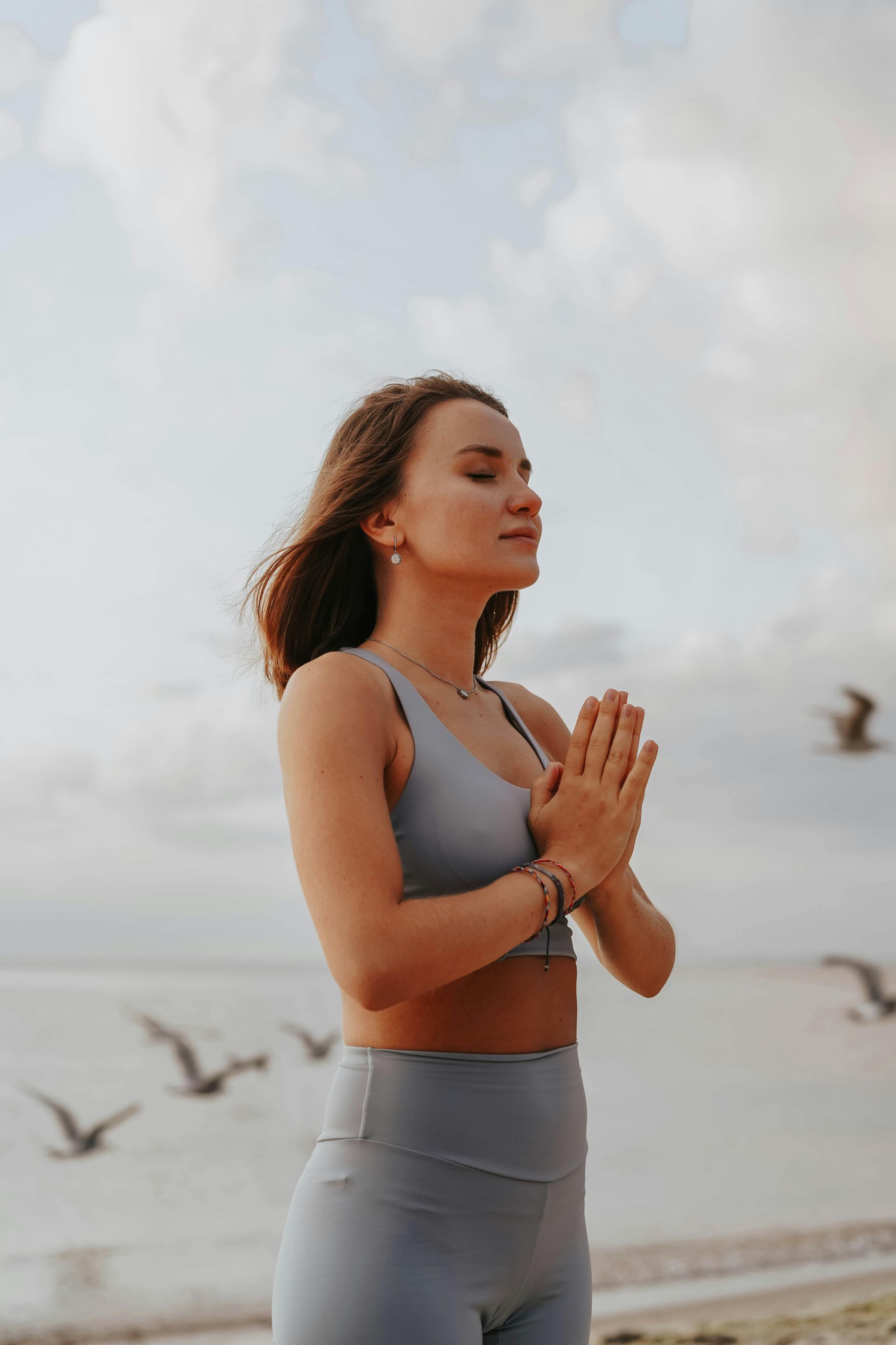 A person in grey athletic wear meditates on a beach with hands pressed together, eyes closed, as birds fly in the sky.