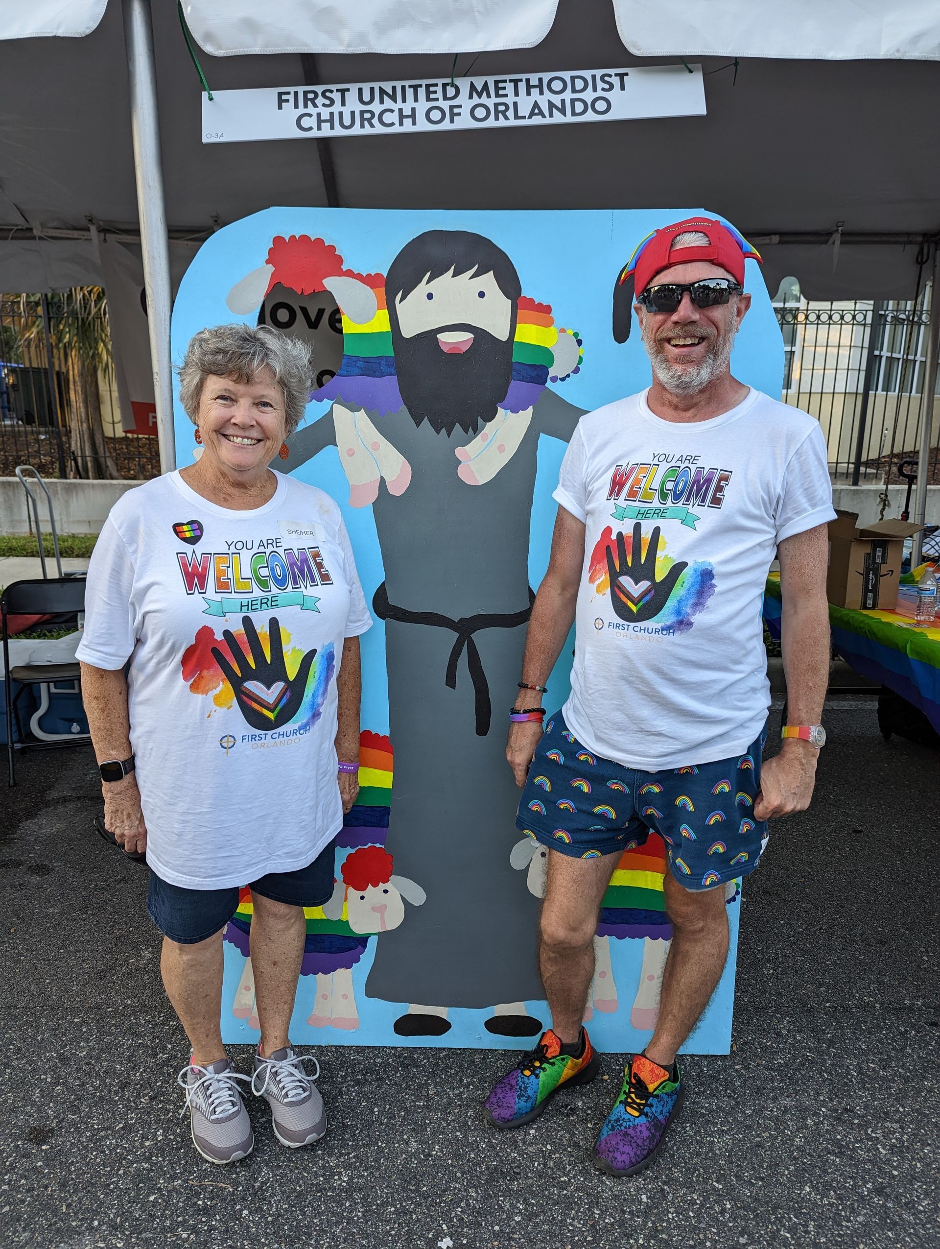 Two people pose with rainbow-framed saint cutout, promoting church at outdoor event.