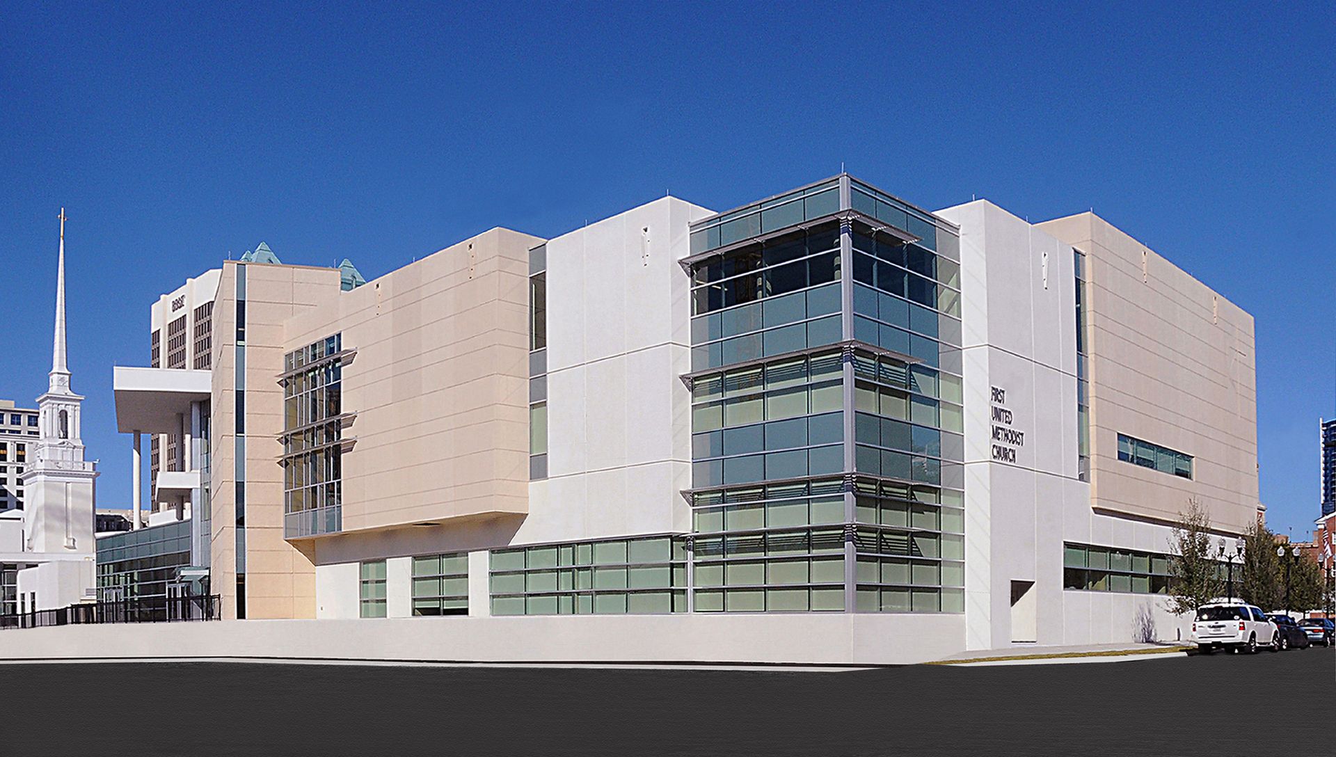 Modern building with beige and white walls, glass tower, and visible windows. Blue sky overhead.