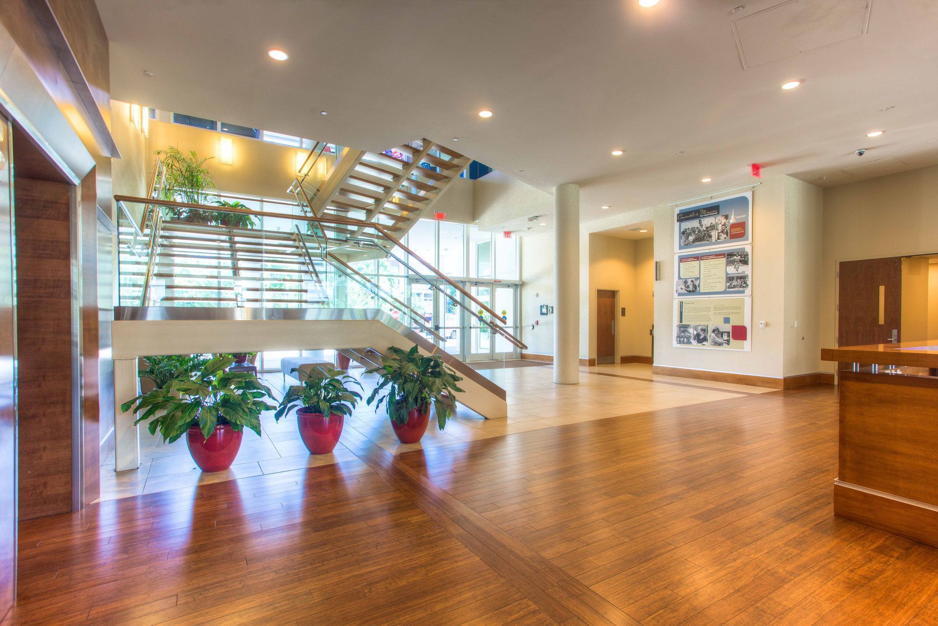 Modern building lobby with wooden floor, staircase, potted plants, and informational display.