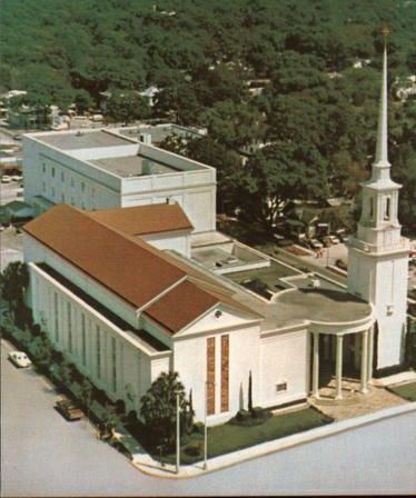Aerial view of a church with a red roof, spire, and colonnade, set against a backdrop of trees.