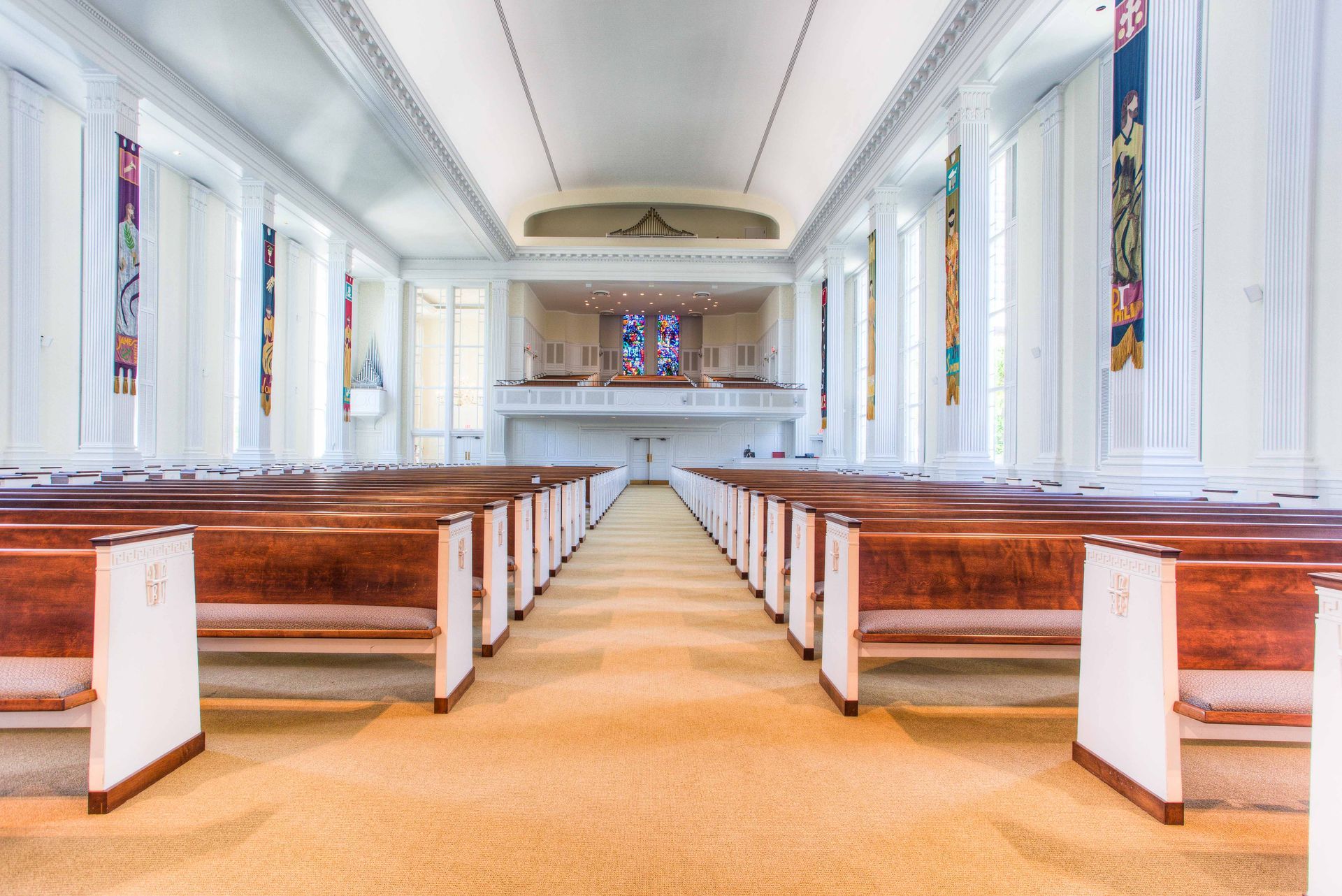 Interior of a light-filled church with rows of wooden pews, white walls, and a stage area.