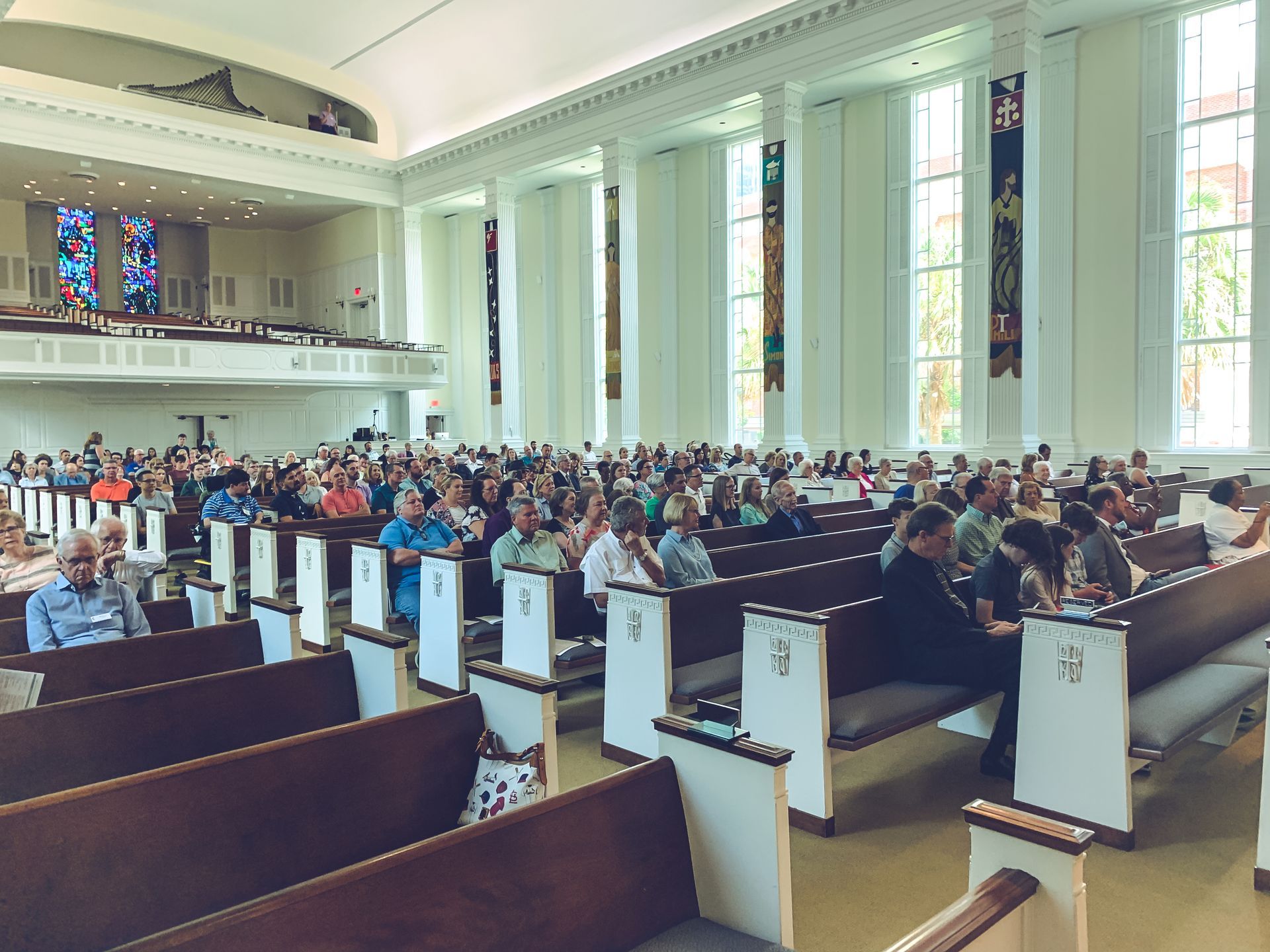 Audience seated in pews inside a large, light-filled church, stained-glass windows visible.