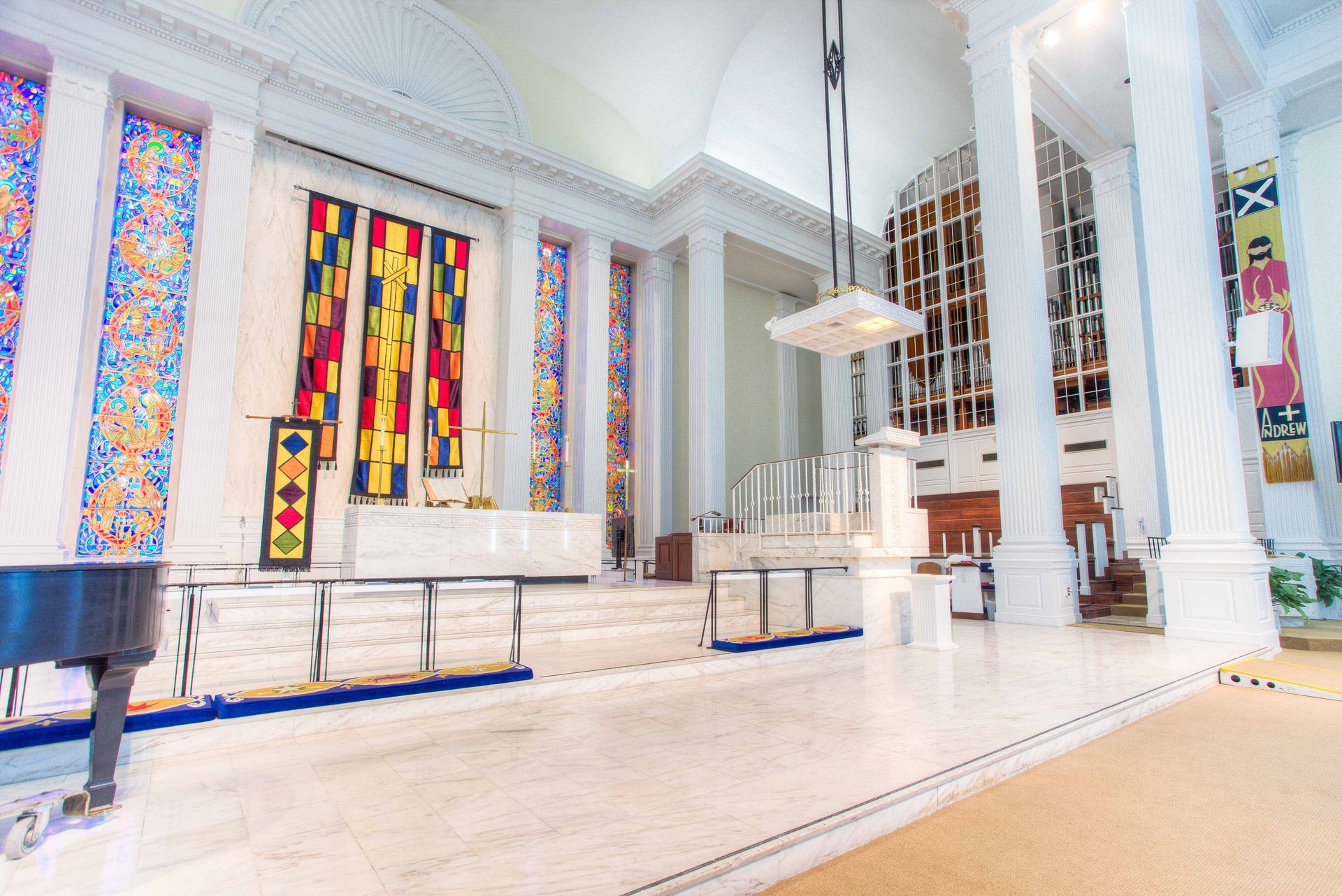 Bright, spacious interior with columns, stained glass windows, and a raised platform. Likely a religious space, possibly a sanctuary.